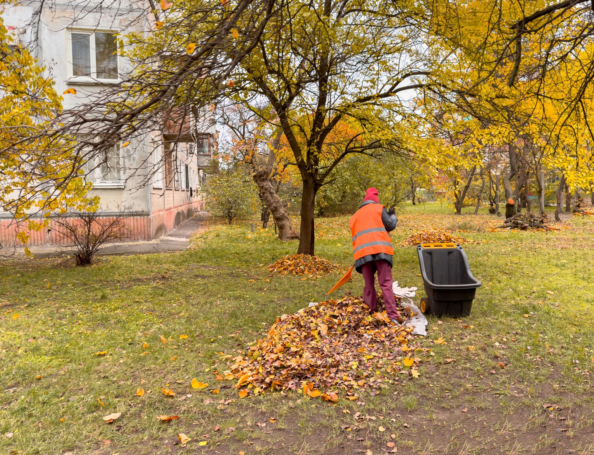 Städtischer Angestellter in Warnweste harkt gelbes Laub in der Nähe eines Wohnhauses. Stadtreinigung im Herbst: Platzwart bei der Arbeit